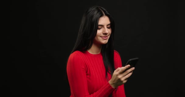 Веселая счастливая брюнетка женщина, использующая смартфон и печатающая изолированные на черном фоне. Улыбнись в камеру с телефоном — стоковое видео