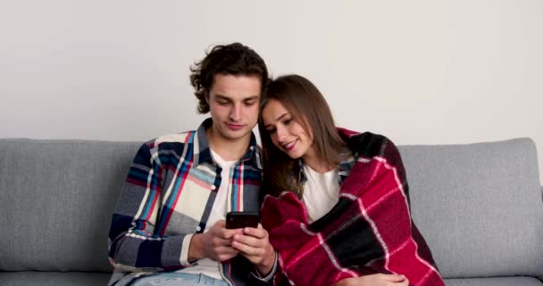 Töprengő pár ül a kanapén, okostelefonokat használnak, mosolyognak és egymásra néznek — Stock videók