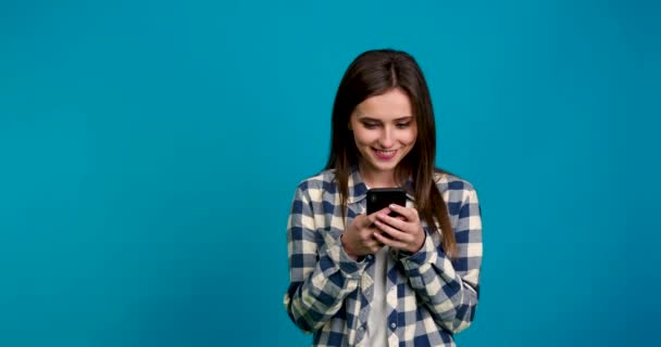 Vista frontal de la chica sonriente en camisa a cuadros usando teléfono inteligente aislado sobre fondo azul — Vídeo de stock