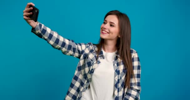 Menina sorridente em camisa quadriculada tomando selfie e enviando beijo de ar isolado no fundo azul — Vídeo de Stock