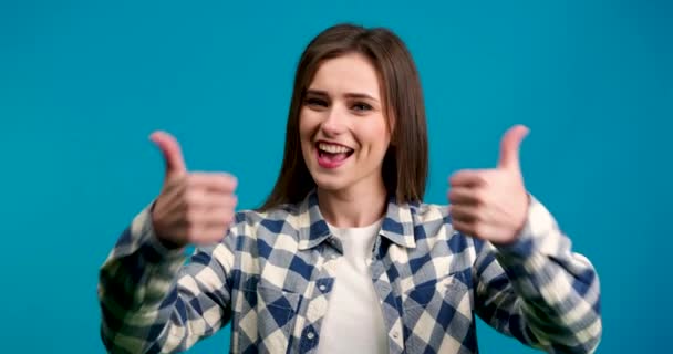 Vooraanzicht van lachend meisje tonen duimen omhoog en kijken naar camera geïsoleerd op blauwe achtergrond — Stockvideo