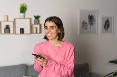 Evde elinde akıllı telefon olan bir kız. Kameraya bak, gülümse.