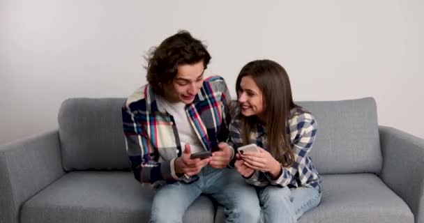 Par som sitter på soffan, ler och använder smartphones i vardagsrummet — Stockvideo