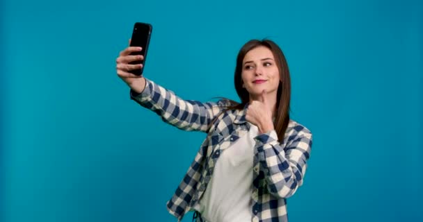 Pige viser fred tegn, tommelfinger op og luft kys, mens du tager selfie isoleret på blå baggrund – Stock-video