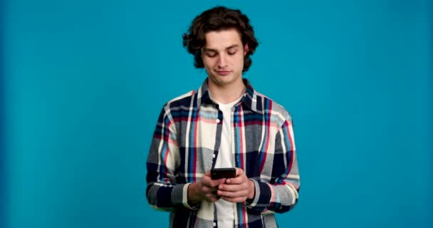 Вид сфокусированного молодого человека, сидящего на смартфоне и улыбающегося на голубом фоне — стоковое видео