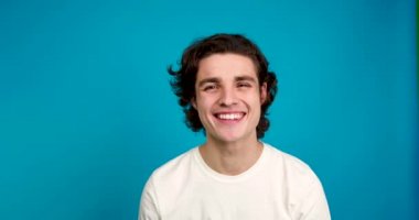 Komik çift saçlarına dokunuyor ve kameraya bakıyor. Mavi arka planda izole edilmiş bir gülümsemeyle.