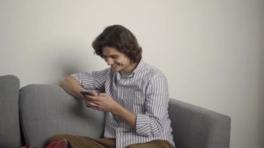Gülümseyen adam kanepede oturuyor ve oturma odasında akıllı telefon kullanıyor.