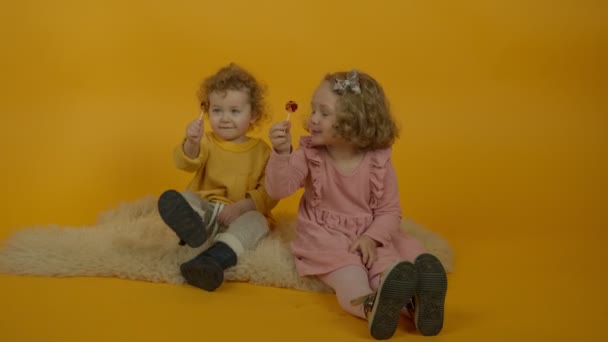 Δύο σγουρά χαμογελαστά παιδιά κάθονται στο χαλί με γλειφιτζούρια — Αρχείο Βίντεο