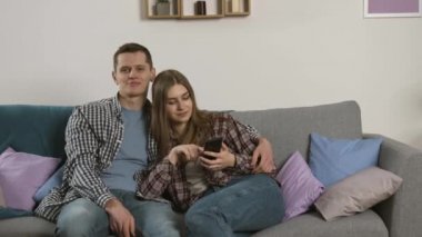 Gülümseyen çift akıllı telefon kullanıyor ve birlikte televizyon izliyorlar.