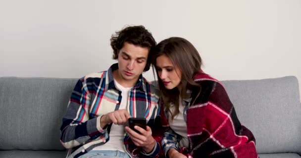 Пара сидит на диване, пользуется смартфонами, улыбается и смотрит друг на друга — стоковое видео