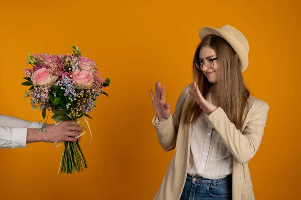 Дівчина в капелюсі і окулярах відмовляється від квітів ізольовано на жовтому тлі — стокове фото