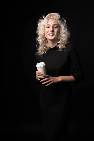 Счастливая блондинка с чашкой кофе, улыбающаяся изолированно на черном фоне — стоковое фото