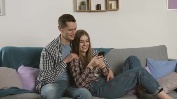 Усміхнена пара в картатих сорочках використовує смартфон на дивані — стокове відео