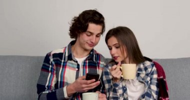 Mutlu çift kanepede oturuyor, çay içiyor ve akıllı telefon kullanıyor.