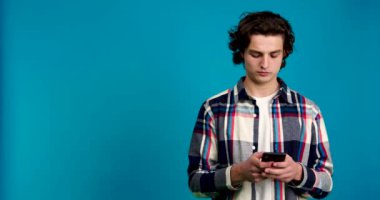 Akıllı telefon kullanan genç adam, mavi arka planda izole edilmiş bir şekilde kafasını sallıyor.