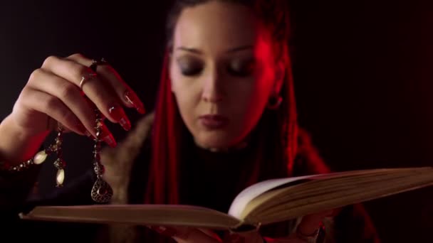 Bruja con amuleto leyendo libro de magia sobre fondo negro — Vídeo de stock