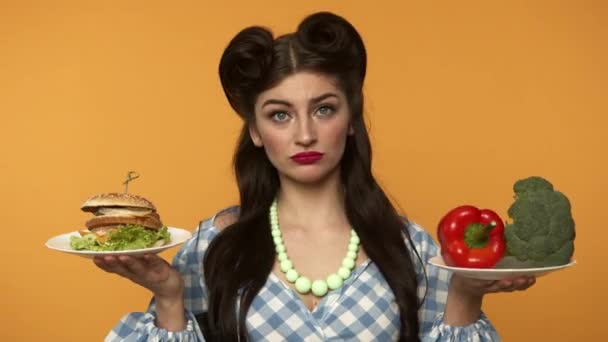 Inseguro pin up mujer sosteniendo hamburguesa y verduras frescas — Vídeo de stock