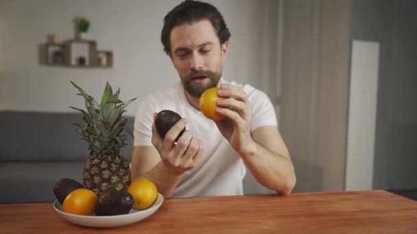 若い男がテーブルに座って果物の有用性について話します,彼はそれらを拾うとパイナップルを表示します,アボカド,オレンジ.果物を中心に — ストック動画