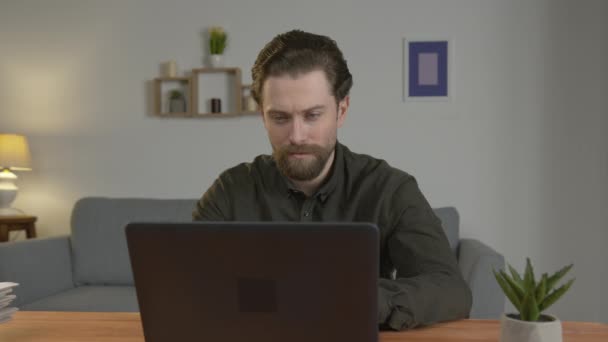 Un hombre blanco con barba se sienta en una mesa, trabaja en un ordenador portátil, trabaja en casa, mira a la cámara, serio . — Vídeo de stock