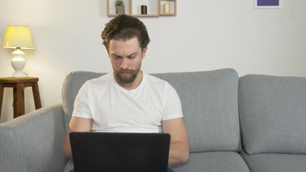 Молодой человек один сидит дома на диване, на своем ноутбуке, ищет работу в Интернете, изолированный от общества из-за карантина — стоковое видео
