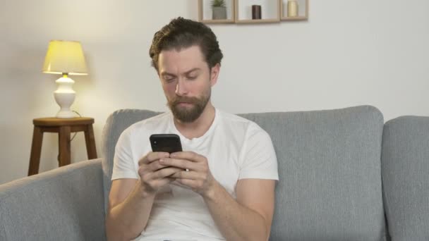 Un jeune homme seul est assis à la maison sur le canapé, communique avec quelqu'un en ligne, isolé, lit les nouvelles — Video