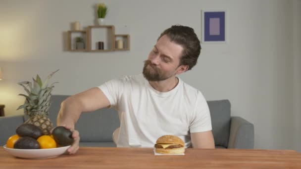 Um cara jovem olha para um prato de frutas e um hambúrguer, pensa, e depois escolhe um hambúrguer, está satisfeito, gosta do sabor de lixo comida ruim . — Vídeo de Stock