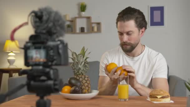 Mladý muž, blogger točí video, sedí u stolu před kamerou a mluví o ovoci a jejich výhodách, pije pomerančový džus. — Stock video