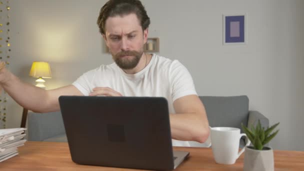 Genç adam evde çalışıyor ve dizüstü bilgisayarla bir masada yemek yiyor, ekrana bakıyor ve kruvasan yiyor, çay içiyor, gülümsüyor. Karantina izolasyonu — Stok video