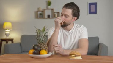 Genç bir adam bir tabak meyveye ve bir hamburgere bakar, meyveyi düşünür ve seçer, sağlıklı bir yaşam tarzı, bir vejetaryen.