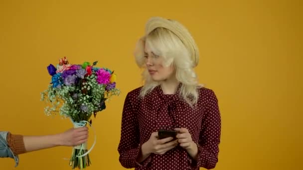 Ξανθιά κοπέλα στο καπέλο χρησιμοποιώντας smartphone και αρνείται μπουκέτο λουλουδιών σε κίτρινο φόντο — Αρχείο Βίντεο