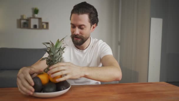 Молодий чоловік сидить за столом і розповідає про корисність фруктів, підбирає їх і показує ананас, авокадо, апельсин. Здорова їжа — стокове відео
