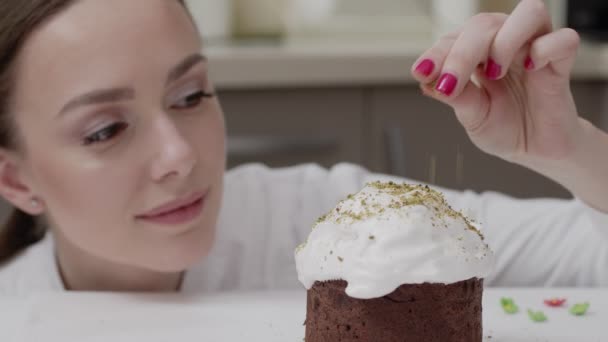 Snoepgoed vrouw in een witte tuniek in een professionele keuken, versiert paastaart, strooit kruimels met pistachenoten, glimlach — Stockvideo