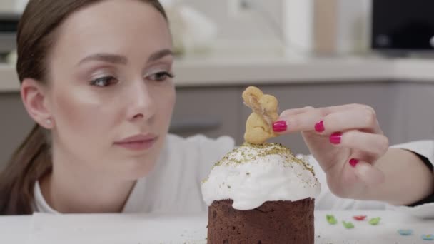 Кондитерша в белой тунике на профессиональной кухне, украшает пасхальный торт, кладет сверху печенье в виде кролика, улыбается — стоковое видео