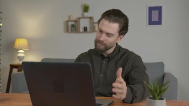 Sakallı beyaz bir adam bir masada oturuyor, internetten iletişim kuruyor, ekrana bakıyor ve esniyor, aceleyle.
