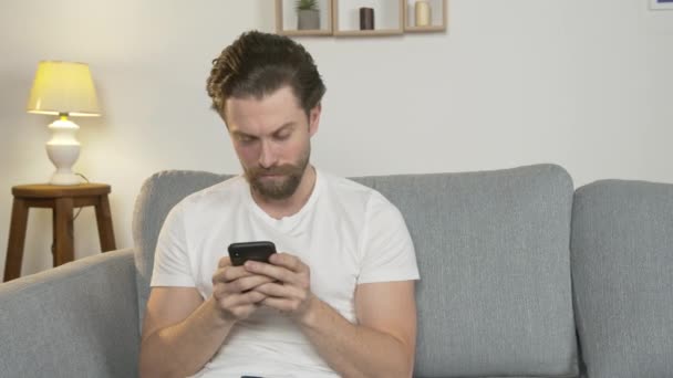 Ein junger Mann sitzt allein zu Hause auf der Couch, schaut auf das Telefon, lächelt, positiv gestimmt, Quarantäne — Stockvideo