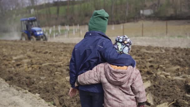 Två pojkar ser hur traktorn odlar marken för plantering av jorden. Plantering av grönsaker för livsmedel — Stockvideo