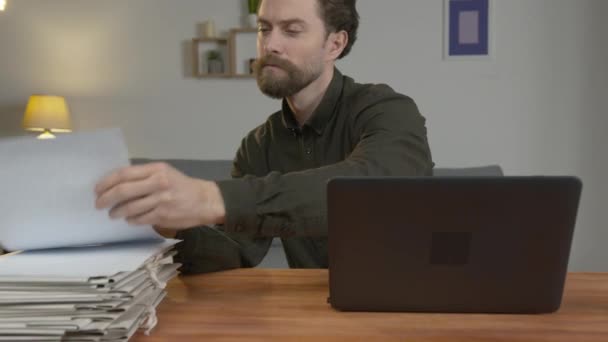 Egy szakállas fehér férfi ül az asztalnál, laptopon dolgozik, otthon dokumentumokkal dolgozik.. — Stock videók