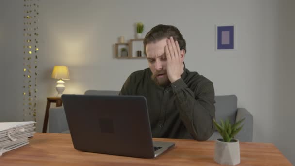 El oficinista con barba se sienta a la mesa con el portátil, mira el monitor del portátil, el hombre es cansado, el trabajo a distancia de casa — Vídeos de Stock