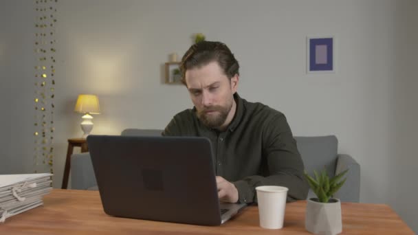 Un bărbat cu barbă stă la o masă, lucrează într-un laptop, acasă, bea cafea fierbinte, este obosit și vrea să doarmă . — Videoclip de stoc