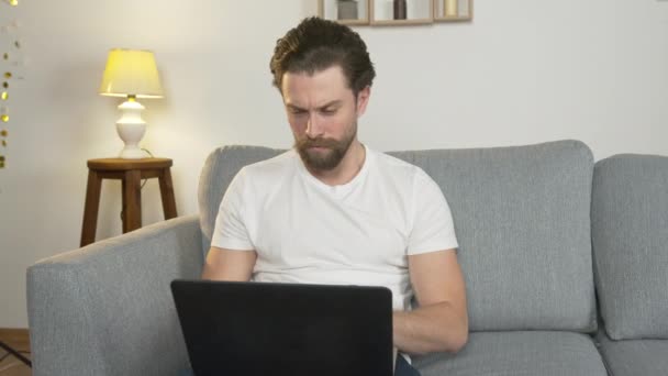 Молодой человек один сидит дома на диване, на коленях ноутбук, серфинг в Интернете, изолирован от общества из-за карантина — стоковое видео