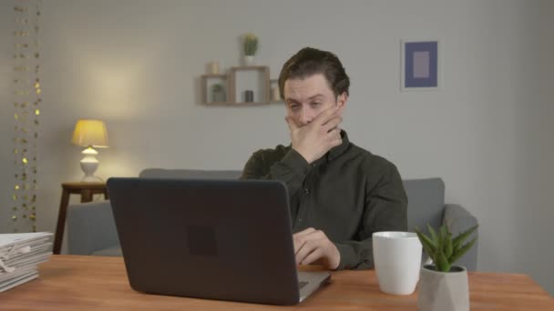 Een blanke man met een baard werkt op een laptop, neemt een doorzichtige fles alcohol en giet het in een witte mok, problemen — Stockvideo