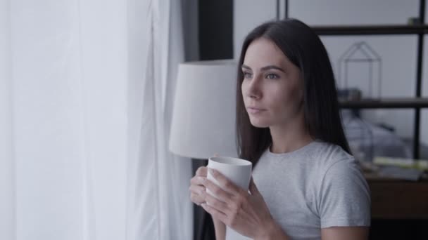 Uma jovem está parada junto à janela bebendo café e olhando para a distância, uma morena delgada com uma xícara nas mãos desfruta do conforto de casa. — Vídeo de Stock