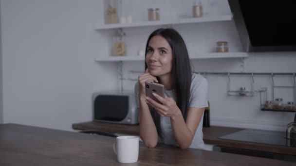 Menina sentada a uma mesa na cozinha, olhando para um smartphone e olhando intensamente para algo, sorrindo, conceito de ficar em casa, compra on-line — Vídeo de Stock