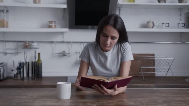 Ein junges Mädchen sitzt an einem Holztisch, liest ein Buch und trinkt ein Getränk aus einer weißen Tasse, das Konzept, Zeit zu Hause zu verbringen, Quarantäne, Freizeit — Stockvideo