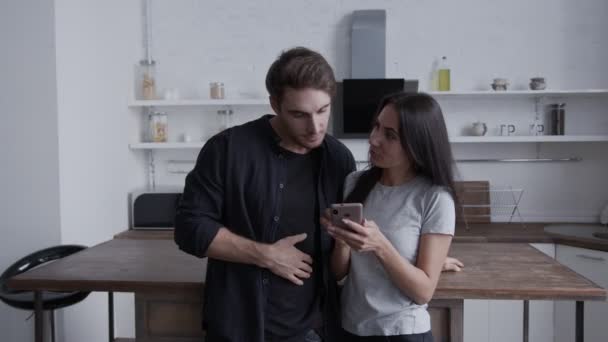 O casal em sua cozinha olhar juntos para o smartphone e se divertir conversando, eles gesticular e sorrir — Vídeo de Stock