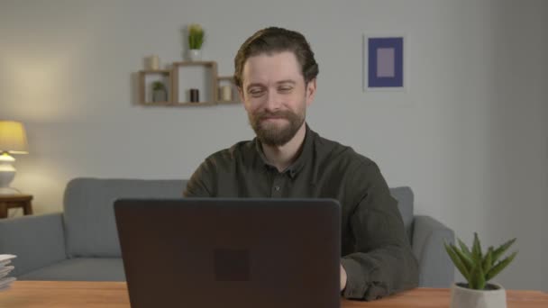 Un hombre blanco con barba se sienta en una habitación en una mesa, se comunica en Internet, mira a su interlocutor a través de una pantalla y agita la mano, sonríe . — Vídeo de stock