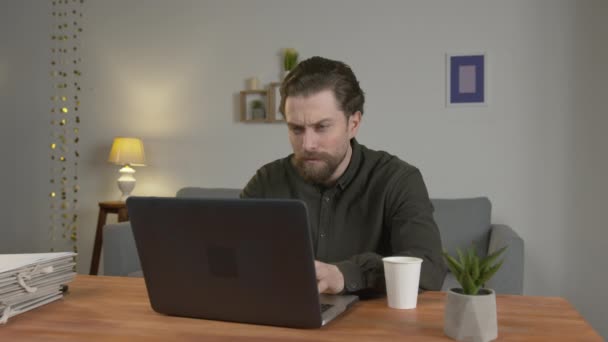 Um homem com uma barba senta-se em uma mesa, trabalha em um laptop, em casa, bebe café quente, ele está cansado e quer dormir . — Vídeo de Stock