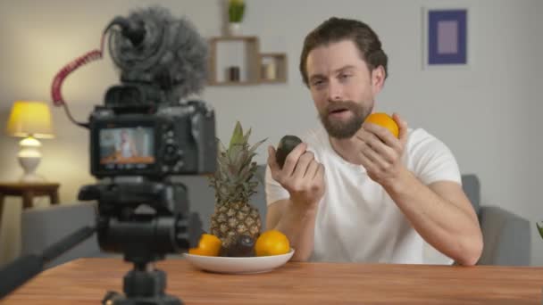 Un joven, vlogger hace un video, se sienta en una mesa frente a la cámara y habla sobre las frutas y sus beneficios — Vídeo de stock