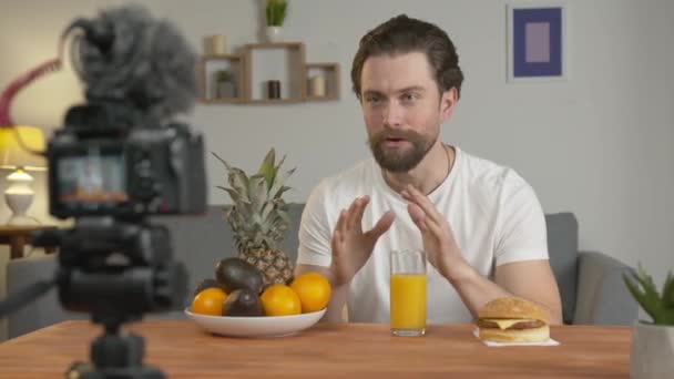 Um jovem, um blogueiro faz um vídeo, senta-se em uma mesa na frente da câmera e fala sobre frutas e seus benefícios, bebe suco de laranja . — Vídeo de Stock