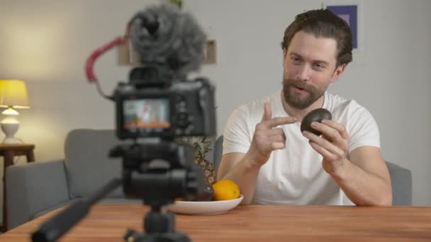 Un giovane, un vlogger fa un video, si siede a un tavolo davanti alla telecamera e parla di frutta e dei loro benefici, l'avocado. — Video Stock